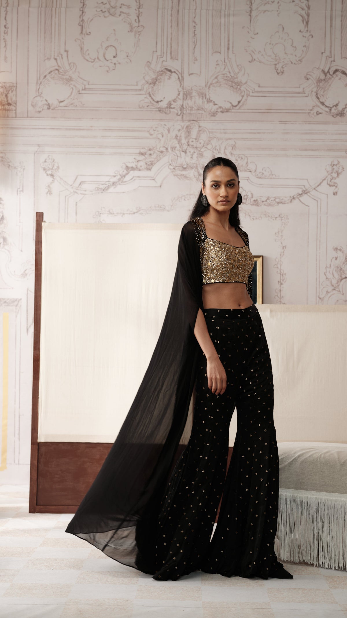 A woman models a black embellished traditional outfit with a long sheer black dupatta, a gold sequined crop top, and black flared pants with gold dot patterns, standing in a classical Indian room with ornate walls and a bed.