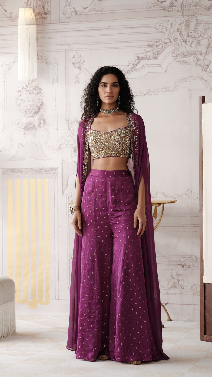A woman stands in a grand, classical-style room wearing a deep purple embroidered traditional outfit with a matching sheer draped overlay, a gold embellished crop top, and high-waisted flared pants.