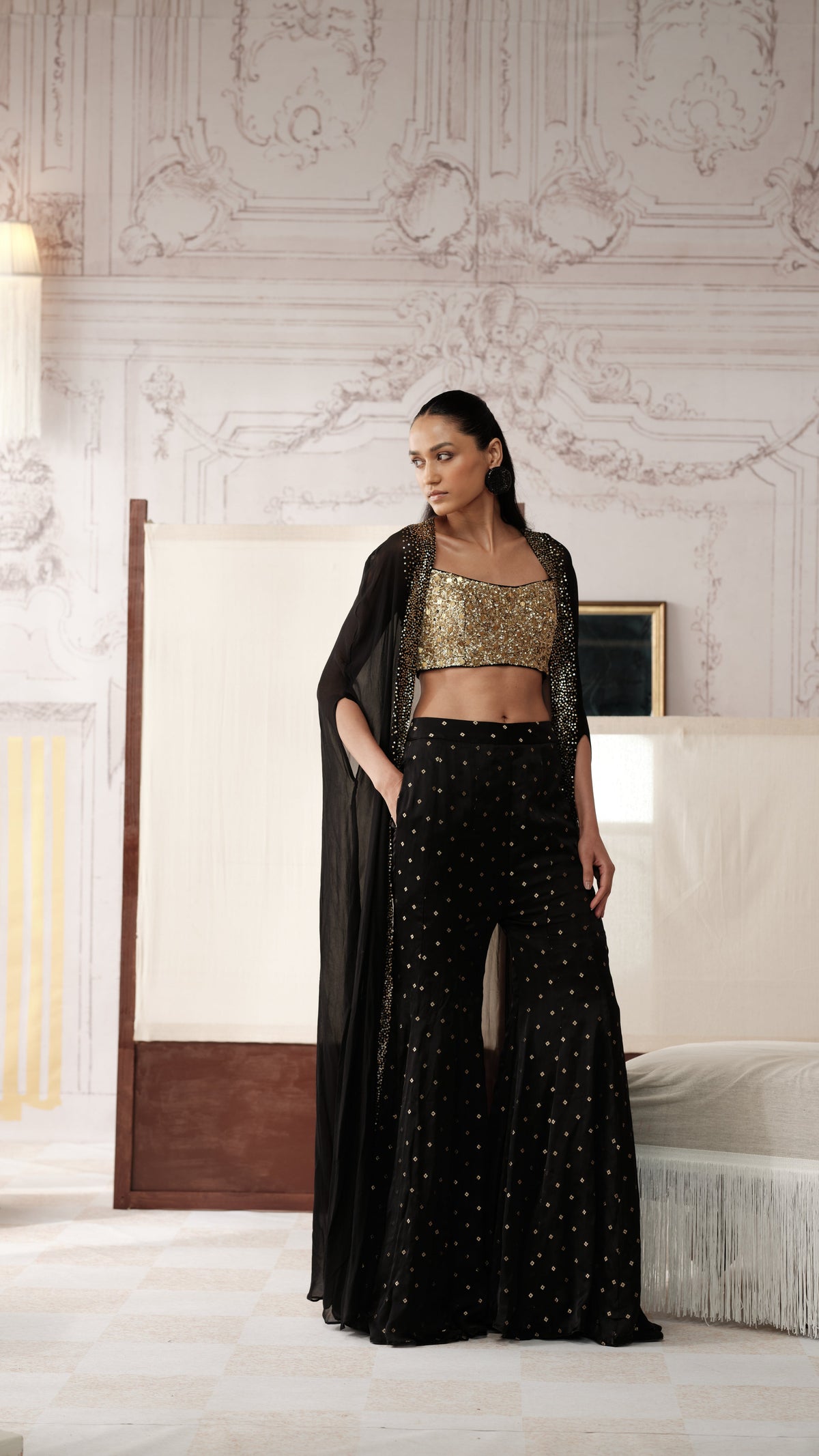 A woman models a black and gold traditional Indian outfit consisting of a gold-embellished crop top, black wide-leg pants with gold dot patterns, and a sheer black dupatta, set against an ornate classical interior background.